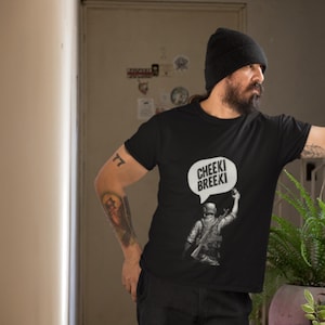 May include: Black t-shirt featuring a graphic of a soldier with a raised arm and a speech bubble that says "Cheeki Breeki" in white. The shirt is worn by a person with a black beanie and a beard.