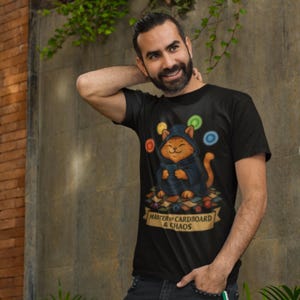 May include: Black t-shirt featuring a cartoon cat wearing a hooded robe and holding a book. The cat is surrounded by colorful circles and sitting on a pile of cardboard. The text "MASTER OF CARDBOARD & CHAOS" is on a banner.