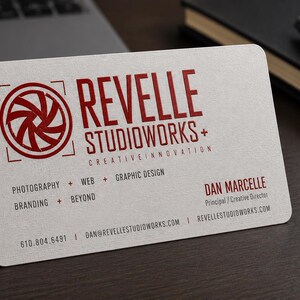 K&ouml;nnte beinhalten: Eine wei&szlig;e Visitenkarte mit abgerundeten Ecken, einem roten Kamera-Blenden-Logo und dem Text REVELLE STUDIOWORKS+ CREATIVE INNOVATION. Sie listet Dienstleistungen wie Fotografie, Web, Grafikdesign, Branding und mehr sowie Kontaktdaten auf.