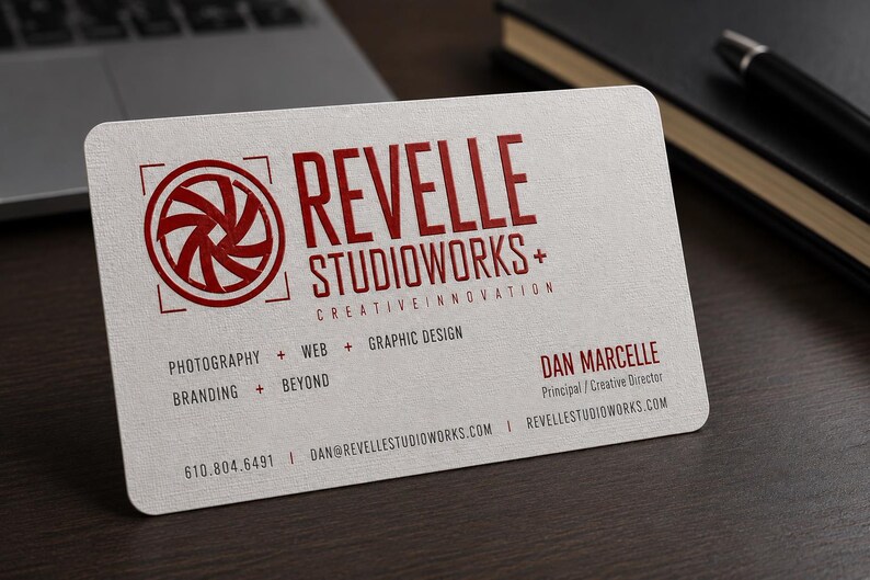 K&ouml;nnte beinhalten: Eine wei&szlig;e Visitenkarte mit abgerundeten Ecken, einem roten Kamera-Blenden-Logo und dem Text REVELLE STUDIOWORKS+ CREATIVE INNOVATION. Sie listet Dienstleistungen wie Fotografie, Web, Grafikdesign, Branding und mehr sowie Kontaktdaten auf.