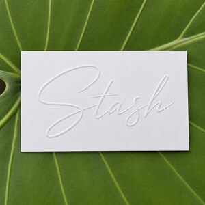 May include: A rectangular white card featuring the word Stash embossed in a cursive script font. The lettering is raised, creating a subtle three-dimensional effect on the smooth surface of the card, which is centered against a green leaf background.