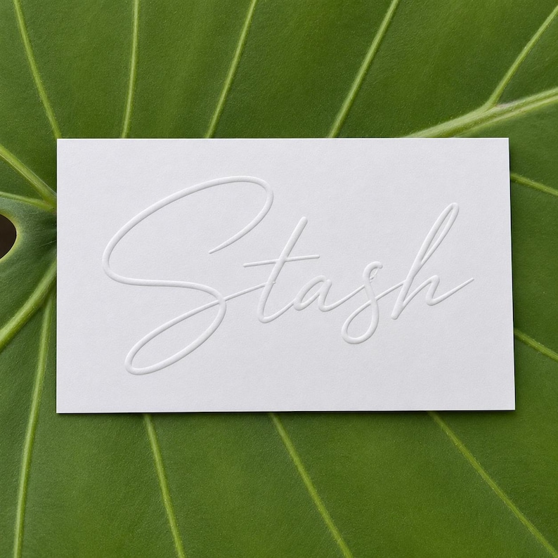 May include: A rectangular white card featuring the word Stash embossed in a cursive script font. The lettering is raised, creating a subtle three-dimensional effect on the smooth surface of the card, which is centered against a green leaf background.