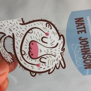 Puede incluir: Una tarjeta de visita rectangular y transparente con esquinas redondeadas que muestra una criatura de dibujos animados con contorno marr&oacute;n, nariz y mejillas rosas. El lado derecho tiene un &aacute;rea azul claro con el texto NATE JOHNSON STUDIOS, natejstudios.com y 703-297-7594.