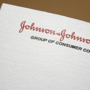 K&ouml;nnte beinhalten: Eine wei&szlig;e, texturierte Karte mit dem roten, kursiven Johnson & Johnson-Logo im oberen Bereich und dem darunter in einer klaren, serifenlosen Schrift aufgedruckten schwarzen Text GROUP OF CONSUMER COMPANIES auf der cremewei&szlig;en Papieroberfl&auml;che.