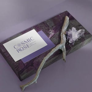 May include: A rectangular purple stone slab featuring a white business card with the text COSMIC ROSE, a weathered grey wooden twig, and a cluster of clear quartz crystals. The card also displays smaller text reading BREATHWORK REIKI MEDITATION.