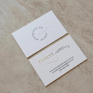 Puede incluir: Dos tarjetas de visita rectangulares blancas. La tarjeta superior presenta un logotipo circular con el texto Evolve Aesthetics Co. La tarjeta inferior muestra el texto Evolve Aesthetics Co. en l&aacute;mina dorada, junto a una web, email y el n&uacute;mero (08) 9203 5382.