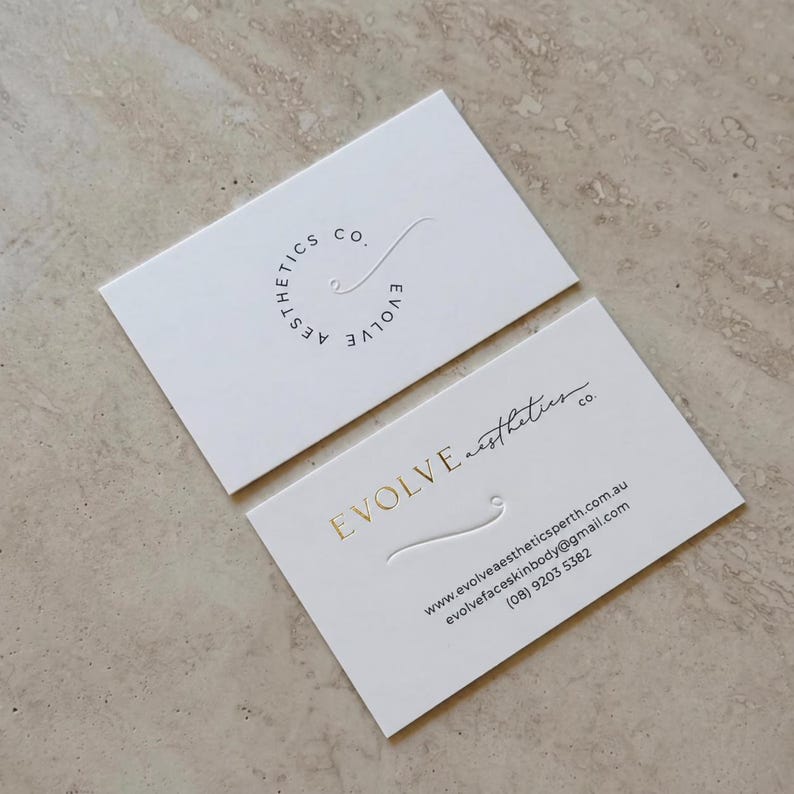 Puede incluir: Dos tarjetas de visita rectangulares blancas. La tarjeta superior presenta un logotipo circular con el texto Evolve Aesthetics Co. La tarjeta inferior muestra el texto Evolve Aesthetics Co. en l&aacute;mina dorada, junto a una web, email y el n&uacute;mero (08) 9203 5382.