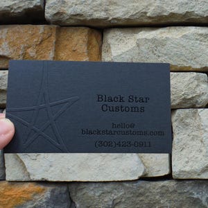 May include: A rectangular black business card featuring an embossed star design on the left side. The text reads Black Star Customs, hello@blackstarcustoms.com, and (302)423-0911. The card has a matte finish with debossed lettering and a star graphic.