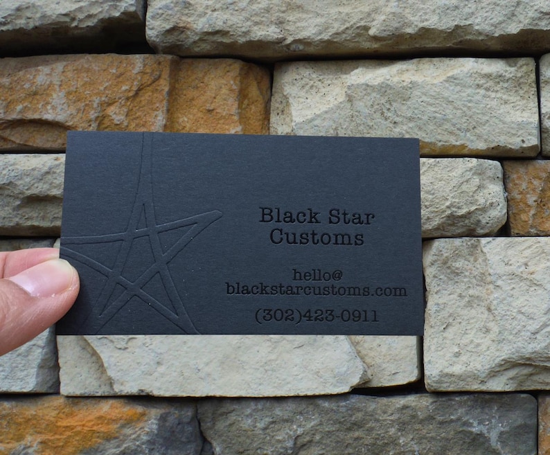 May include: A rectangular black business card featuring an embossed star design on the left side. The text reads Black Star Customs, hello@blackstarcustoms.com, and (302)423-0911. The card has a matte finish with debossed lettering and a star graphic.