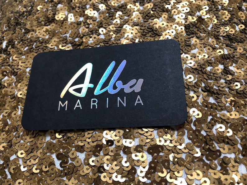 May include: A rectangular black business card with rounded corners featuring the text Alba in a holographic script font and the word MARINA in a white sans-serif font, positioned against a background of shimmering gold sequins.