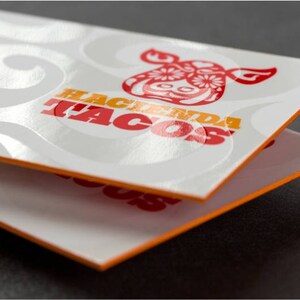May include: Two white business cards with orange edges stacked on top of each other. The top card has a white background with a red and yellow logo that says "Hacienda Tacos" with a stylized red cow head design. The logo is embossed and has a glossy finish.