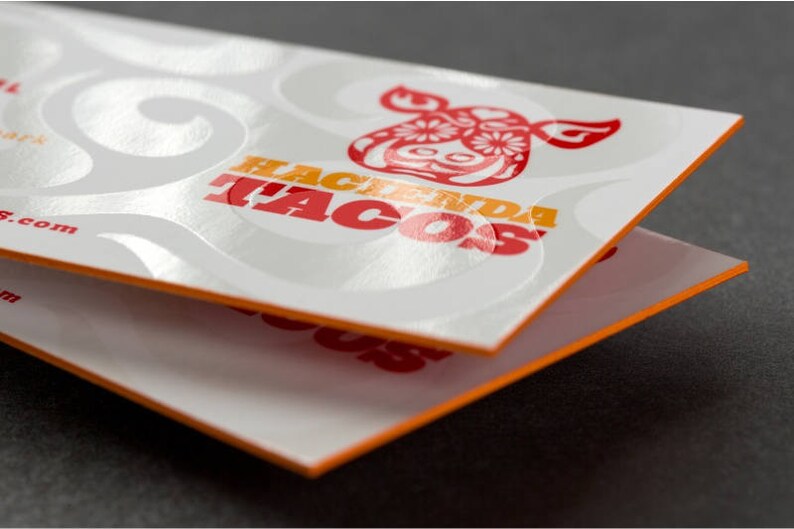 May include: Two white business cards with orange edges stacked on top of each other. The top card has a white background with a red and yellow logo that says "Hacienda Tacos" with a stylized red cow head design. The logo is embossed and has a glossy finish.