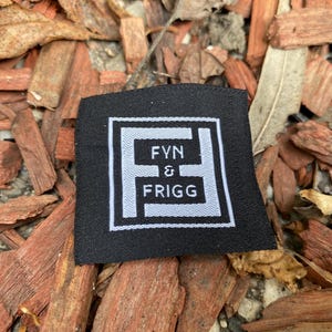 May include: A square black fabric clothing label featuring a white embroidered border and a central white logo. The logo consists of a stylized letter F framing the text FYN & FRIGG in white capital letters, centered on the black background.