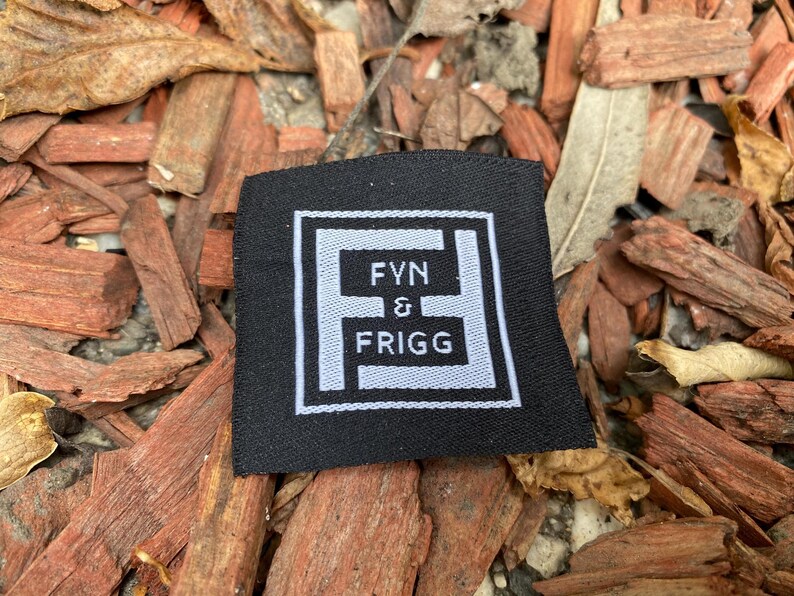 May include: A square black fabric clothing label featuring a white embroidered border and a central white logo. The logo consists of a stylized letter F framing the text FYN & FRIGG in white capital letters, centered on the black background.