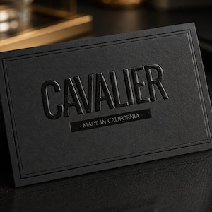 May include: A rectangular black business card featuring the word CAVALIER in large, embossed, glossy black lettering. Below the main text, a smaller rectangular area contains the words MADE IN CALIFORNIA. The card has a double-line border.