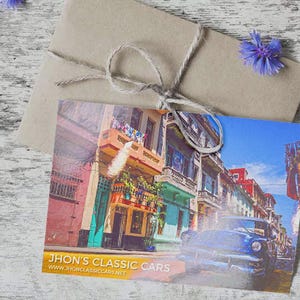 May include: A rectangular postcard featuring a vibrant street scene with colorful buildings and a blue vintage car, with the text "JHON'S CLASSIC CARS" and "WWW.JHONCLASSICCARS.NET" printed at the bottom, resting on a brown paper envelope tied with twine.