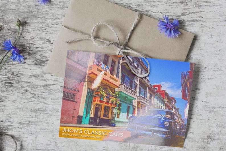 May include: A rectangular postcard featuring a vibrant street scene with colorful buildings and a blue vintage car, with the text "JHON'S CLASSIC CARS" and "WWW.JHONCLASSICCARS.NET" printed at the bottom, resting on a brown paper envelope tied with twine.