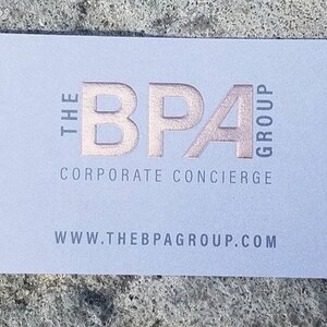 Puede incluir: Una tarjeta de visita rectangular con fondo gris claro. El texto THE BPA GROUP est&aacute; impreso en l&aacute;mina de oro rosa met&aacute;lico. Debajo, las palabras CORPORATE CONCIERGE y el sitio web WWW.THEBPAGROUP.COM est&aacute;n impresas en gris oscuro.