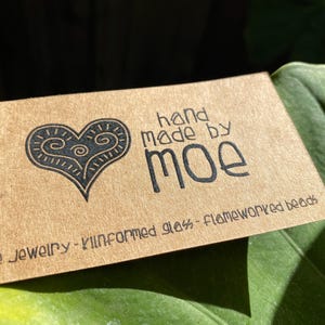May include: A rectangular tan card with black text and a heart graphic. The text reads hand made by moe, fine jewellery - kilnformed glass - flameworked beads. The heart graphic features internal swirl patterns and a pointed base.