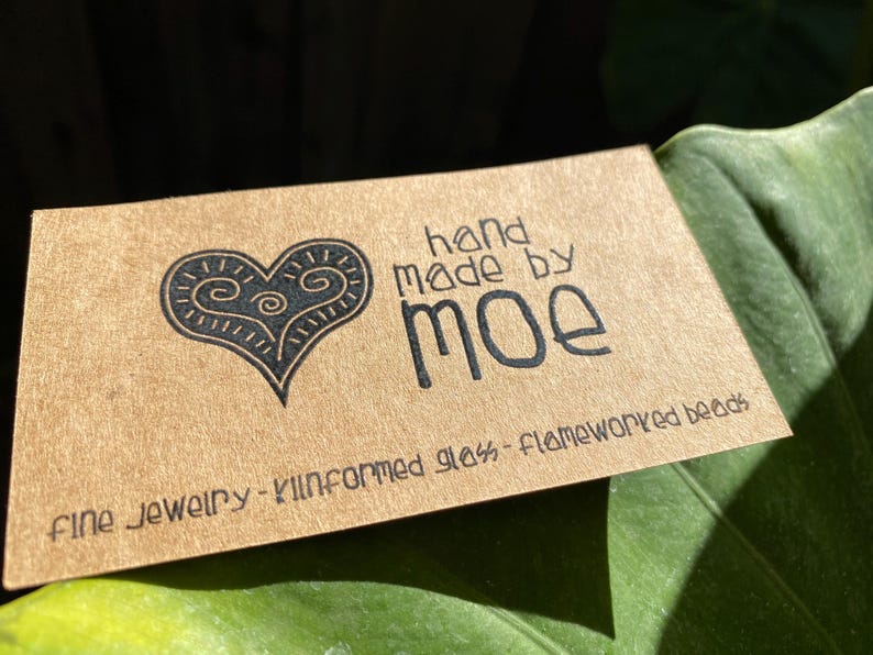 May include: A rectangular tan card with black text and a heart graphic. The text reads hand made by moe, fine jewellery - kilnformed glass - flameworked beads. The heart graphic features internal swirl patterns and a pointed base.