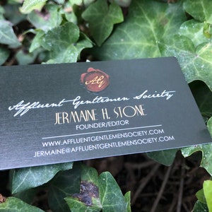 May include: A black business card with gold lettering. The card reads "Affluent Gentlemen Society" and "Jermaine H. Stone, Founder/Editor". The card also includes the website address "www.affluentgentlemensociety.com" and the email address "jermaine@affluentgentlemensociety.com".