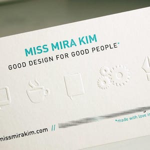 May include: A white business card with the text "MISS MIRA KIM" in teal, followed by "GOOD DESIGN FOR GOOD PEOPLE." Embossed icons include a computer monitor, coffee cup, notebook, gears, and a pen nib. The website address is visible at the bottom.