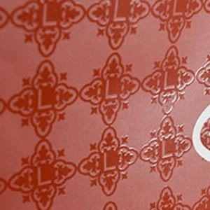 May include: A vibrant red surface featuring a repeating pattern of dark red, stylized cross-shaped motifs, each containing the letter L. A large, white, ornate, symmetrical emblem featuring a central letter L is positioned on the right side.