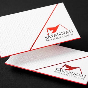 May include: Two white rectangular business cards with red edges. The cards feature a textured geometric pattern on the left and a red logo with the text SAVANNAH REAL ESTATE COMPANY on the right, separated by a diagonal red line.