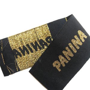 May include: A black fabric label featuring the word PANINA in gold lettering. One section of the label displays the text in gold embroidery on a black background, while the other section features the word PANINA in black on a gold metallic background.