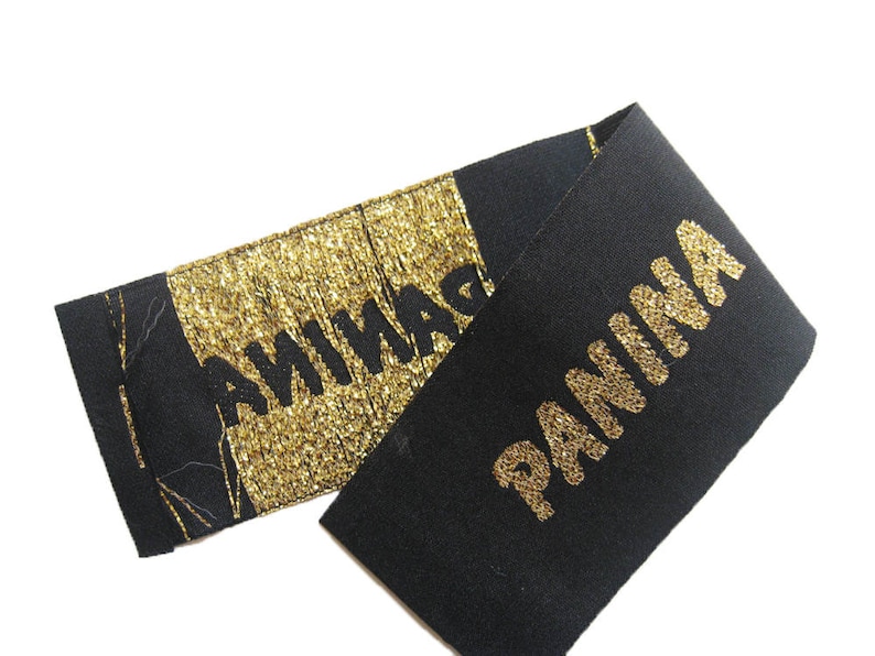 May include: A black fabric label featuring the word PANINA in gold lettering. One section of the label displays the text in gold embroidery on a black background, while the other section features the word PANINA in black on a gold metallic background.