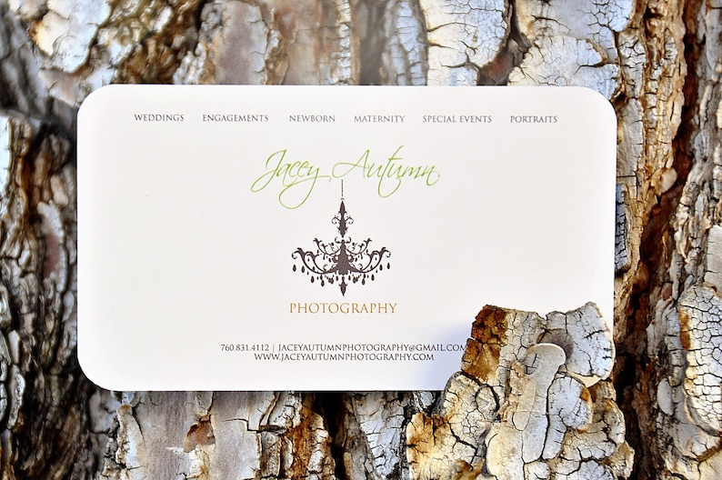 May include: A rectangular white business card with rounded corners featuring the text Jacey Autumn Photography in green and gold script and print. A dark brown chandelier graphic is centered. Text includes services, phone number, email, and website.