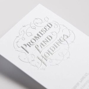 Puede incluir: Una tarjeta de visita blanca con texto en relieve de l&aacute;mina plateada que dice Promised Land Holdings, rodeado por un borde circular decorativo de alambre de espino. La tarjeta tiene un dise&ntilde;o limpio con letras plateadas met&aacute;licas.