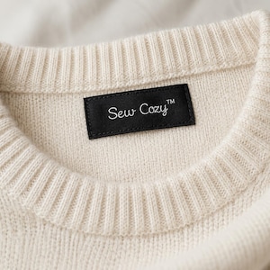 May include: A cream-colored knit sweater featuring a ribbed crew neckline and a rectangular black fabric label sewn into the inner collar. The label displays the white text Sew Cozy with a trademark symbol in a simple, clean font style.