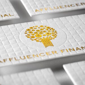 May include: A white business card with a grid-like embossed texture. The card features a gold foil logo of a tree made of circular coin shapes and the text AFFLUENCER FINANCIAL printed in gold foil at the bottom of the card surface.