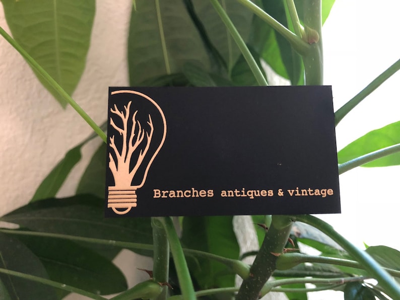 May include: A rectangular black business card featuring a gold-colored graphic of a lightbulb with tree branches inside the glass bulb. The text "Branches antiques & vintage" is printed in gold lettering on the right side of the card.