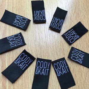May include: A collection of nine rectangular black fabric labels arranged in a circular pattern. Each label features the text CODY & KAIT embroidered in white thread in a clean, sans-serif font, oriented vertically across the center of the fabric.