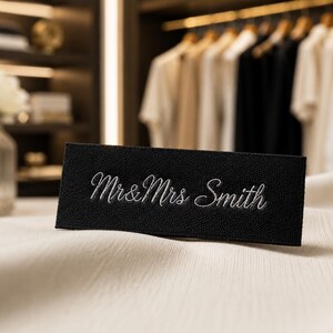 May include: A rectangular black fabric label featuring the embroidered white text Mr & Mrs Smith in a cursive script font. The label is positioned on a light-colored, textured fabric surface, displaying clear stitching and a clean, finished edge.