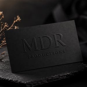May include: A rectangular black business card featuring the embossed text MDR PRODUCTIONS in a serif font. The card has a matte finish and is positioned on a dark, textured surface next to a small, dried, light-colored botanical branch.