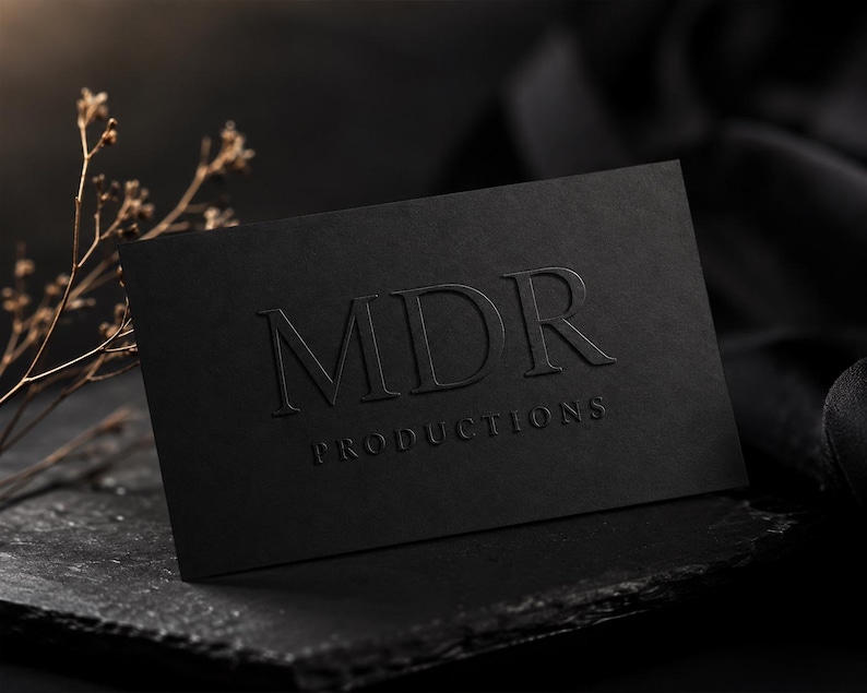May include: A rectangular black business card featuring the embossed text MDR PRODUCTIONS in a serif font. The card has a matte finish and is positioned on a dark, textured surface next to a small, dried, light-colored botanical branch.