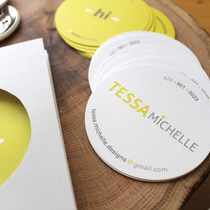 Puede incluir: Una pila de tarjetas de visita circulares blancas con el texto amarillo TESSA MICHELLE, un n&uacute;mero de tel&eacute;fono y una direcci&oacute;n de correo electr&oacute;nico. Al lado hay una tarjeta blanca con un recorte circular que revela un fondo amarillo con el texto gris hi.