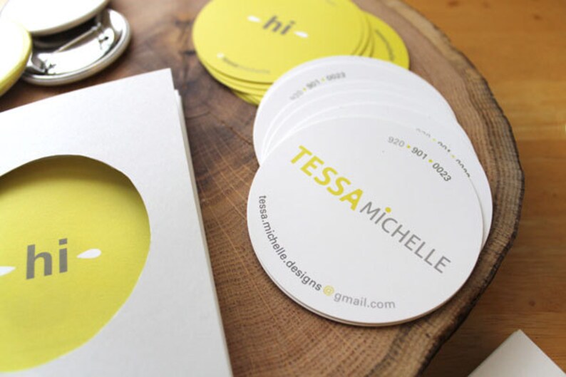 Puede incluir: Una pila de tarjetas de visita circulares blancas con el texto amarillo TESSA MICHELLE, un n&uacute;mero de tel&eacute;fono y una direcci&oacute;n de correo electr&oacute;nico. Al lado hay una tarjeta blanca con un recorte circular que revela un fondo amarillo con el texto gris hi.