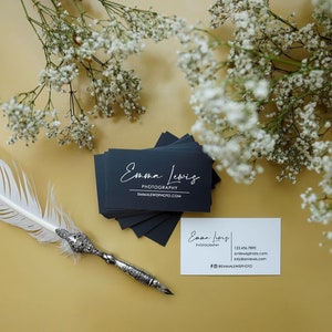 May include: A stack of rectangular black business cards with white text reading Emma Lewis Photography and emmalewisphoto.com, alongside a single white business card with contact details and a white feather quill pen with an ornate silver handle.