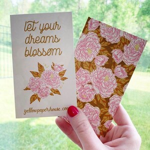 May include: Two rectangular paper cards. One card is cream-colored with the gold text 'let your dreams blossom', a floral illustration of pink peonies with gold leaves, and 'yellowpaperhouse.com'. The other card features a repeating pink peony pattern.