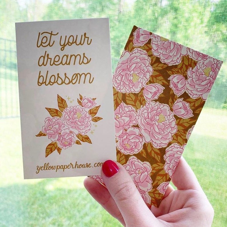 May include: Two rectangular paper cards. One card is cream-colored with the gold text 'let your dreams blossom', a floral illustration of pink peonies with gold leaves, and 'yellowpaperhouse.com'. The other card features a repeating pink peony pattern.