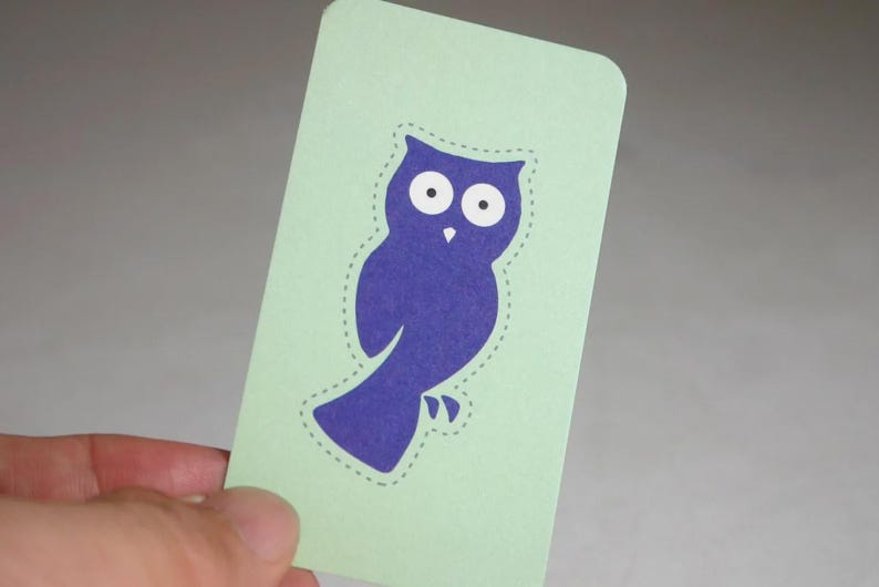 May include: A rectangular light green card featuring a dark purple owl silhouette in the center. The owl has two large white circular eyes with black pupils and a small white beak. A dashed line outlines the shape of the owl on the card.