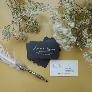 May include: A stack of dark charcoal business cards with the white text Emma Lewis Photography and emmalewisphoto.com, alongside a single white business card with contact details, a silver quill pen, and sprigs of white baby's breath flowers.