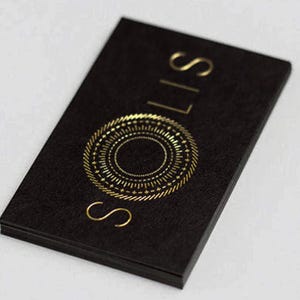 May include: A rectangular black business card with a textured surface. The card features the word SOLIS printed in gold foil lettering, arranged vertically. Below the text is a circular gold foil graphic resembling a sun with radiating lines.