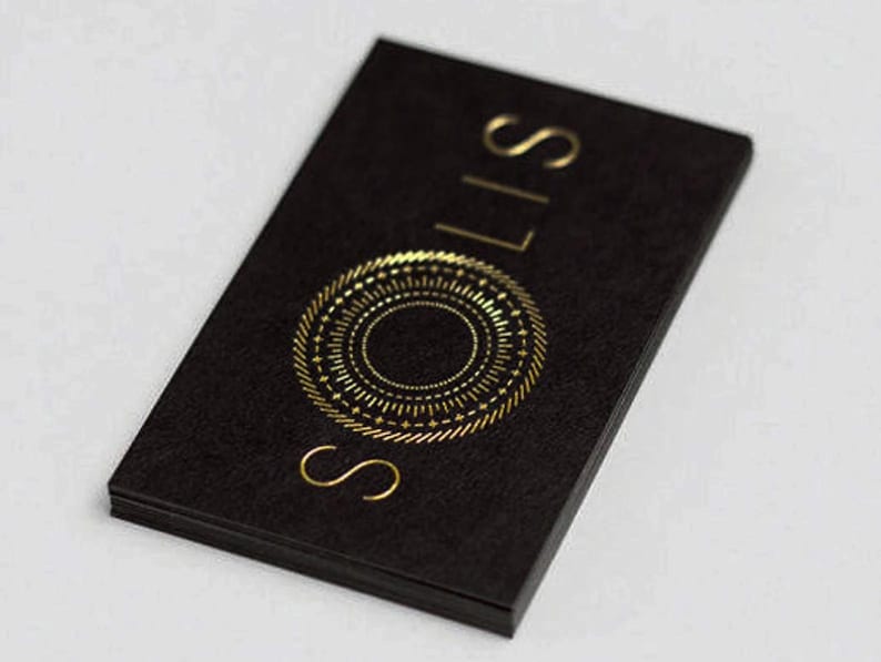 May include: A rectangular black business card with a textured surface. The card features the word SOLIS printed in gold foil lettering, arranged vertically. Below the text is a circular gold foil graphic resembling a sun with radiating lines.