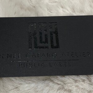 May include: A rectangular black business card featuring a glossy embossed logo at the top. Below the logo, the text reads RENEE CAFARO ATELIER with a horizontal line underneath, followed by the text PUBLIC LABEL in a clean, sans-serif font.