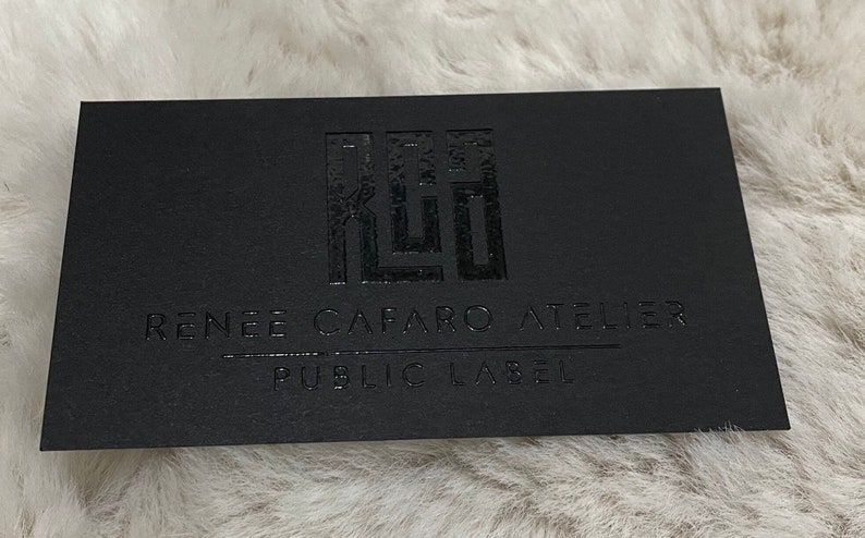 May include: A rectangular black business card featuring a glossy embossed logo at the top. Below the logo, the text reads RENEE CAFARO ATELIER with a horizontal line underneath, followed by the text PUBLIC LABEL in a clean, sans-serif font.
